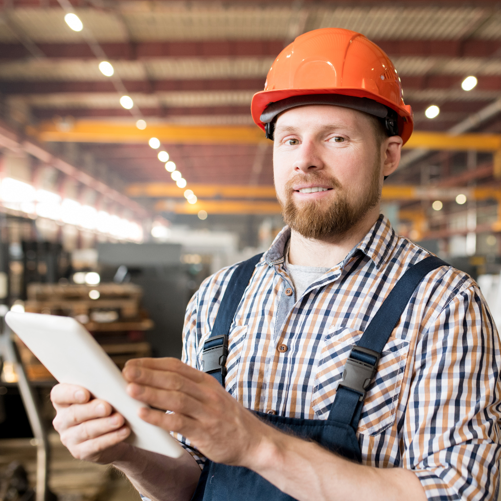 Homem em ambiente industrial usando capacete laranja e segurando um tablet.